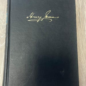 May include: A black hardcover book with the name Henry James embossed in gold script on the front cover. The book features a vertical spine on the left side and a rectangular shape with a solid dark fabric texture covering the front surface.