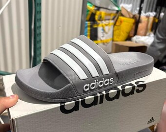 May include: A grey Adidas slide sandal featuring three white stripes across the top strap and the Adidas logo printed in white on the side. The sandal is resting on top of a white Adidas shoebox with the brand name printed in large black letters.