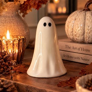 May include: A smooth, white ceramic ghost figurine with a rounded top and a flared base. The ghost features two simple, solid black oval eyes positioned near the top. The surface of the figurine is glossy and reflects light, showing subtle vertical folds.