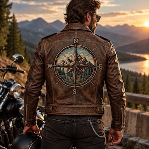 May include: A brown leather jacket featuring a large embroidered compass rose design on the back. The embroidery depicts a mountain landscape with pine trees and the cardinal directions N, S, E, and W. The jacket has long sleeves with zippered cuffs.