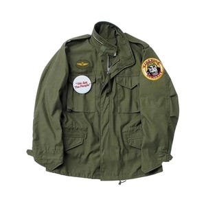May include: An olive green field jacket featuring a high collar, a front zipper, and four flap pockets. It includes a gold-toned wing patch, a round white button with the text "We Are The People," and a circular "King Kong Company" patch.