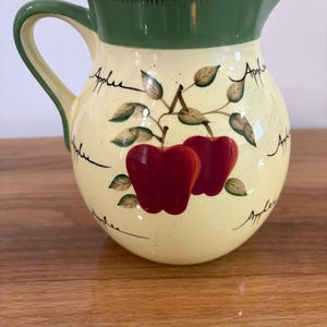 May include: A ceramic pitcher with a cream-colored body and a green rim. The pitcher features a painted design of two red apples with green leaves and the word Apples written in black script multiple times around the exterior surface.