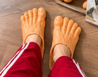 May include: A pair of slip-on slippers shaped like large, realistic human feet. The slippers are light tan in color, featuring molded toes, detailed skin texture, and raised vein patterns across the top surface of each foot-shaped shoe.