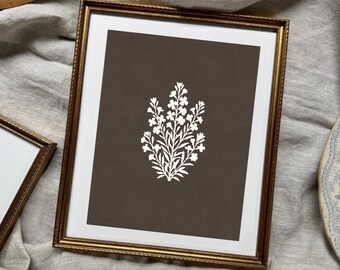 May include: A rectangular wall art piece featuring a white botanical illustration of a flowering plant centered on a dark brown background, enclosed within a thin, textured gold-toned frame with a white mat border surrounding the central artwork.