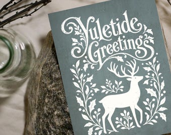May include: A rectangular greeting card with a muted blue-gray background featuring the white text Yuletide Greetings above a white illustration of a deer surrounded by a decorative border of holly leaves and berries on a textured surface.