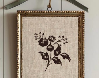 May include: A rectangular wall hanging featuring a dark brown floral illustration printed on a beige burlap fabric background. The piece is encased in a metallic gold frame with a beaded edge and is suspended by a jute cord and a sage green ribbon bow.