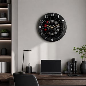 May include: A circular wall clock designed to resemble a black car wheel rim with a tyre tread outer edge. The clock face features white Arabic numerals from one to twelve, gold-coloured hour and minute hands, and a visible red brake caliper.