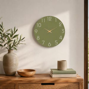 Peut inclure: Une horloge murale ronde au cadran vert olive mat, dotée de chiffres blancs fins de 1 à 12 et d'aiguilles dorées fines pour les heures et les minutes. L'horloge présente un design circulaire épuré sans cadre, offrant une esthétique simple et fonctionnelle.