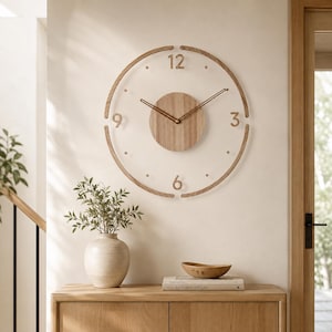 May include: A circular wall clock featuring a transparent face with a light wood centre disc, a thin wood outer ring, and wood numerals at the 12, 3, 6, and 9 positions. The clock has two thin, gold-toned metal hands centred on the wood disc.