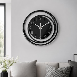 Könnte beinhalten: Eine runde Wanduhr mit schwarzem Zifferblatt, weißen arabischen Ziffern und dünnen weißen Zeigern. Die Uhr ist von zwei konzentrischen schwarzen Ringen umgeben, die ein offenes, geschichtetes Design bilden und den Blick auf die Wand freigeben.