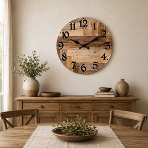 Könnte beinhalten: Eine große, runde Wanduhr aus horizontalen Holzplanken in verschiedenen hellen und dunklen Brauntönen. Das Zifferblatt zeigt kräftige schwarze arabische Ziffern von eins bis zwölf sowie zwei schwarze Metallzeiger in der Mitte der Uhr.