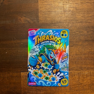 May include: A holographic trading card featuring Thrasios Triton Hero, a blue merfolk wizard eating cereal with green and blue mana symbols. The card includes text: Partner Up For Bigger Plays, Mana Value, Legendary Creature Merfolk Wizard, Scry Draw Repeat, and 1/3.