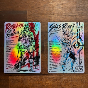 May include: Two rectangular trading cards with a holographic finish. The left card features Rograkh, Son of Rohgahh, a legendary creature with red ink splatters. The right card features Silas Renn, Seeker Adept, a legendary creature with blue ink splatters.