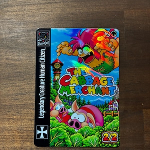May include: A rectangular trading card with a holographic finish featuring a cartoon character and two pigs in a forest. The text reads THE CABBAGE MERCHANT, Legendary Creature Human Citizen, and 2/2. A CrusaderCraft logo is in the top left corner.