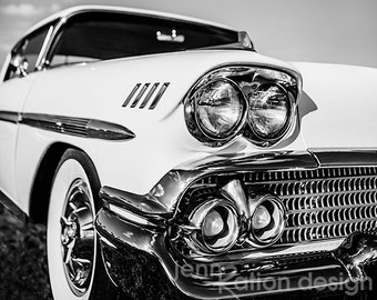 May include: Black and white photo of a classic white car with chrome accents. The car has a large chrome grille and two sets of headlights. The car is parked in front of a cloudy sky.