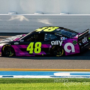 Peut inclure: Une voiture de course noire et rose avec le numéro 48 en vert néon. La voiture porte le logo Ally et la marque Microsoft. La voiture est sur un circuit avec une bande bleue et jaune.