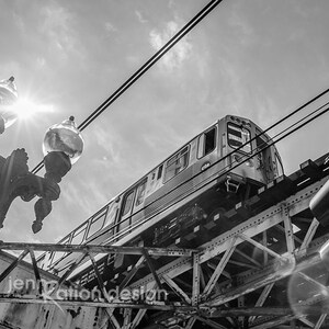 May include: A black and white photo of a train traveling over a bridge. The train is in the foreground and the bridge is in the background. The train is a silver color and has windows on both sides. The bridge is made of metal and has a lot of beams. The sky is cloudy and there is a streetlight in the foreground.