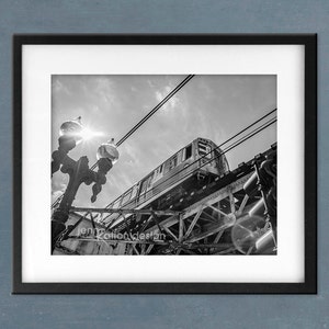 May include: A black and white photograph of a train passing under a bridge. The train is seen from a low angle, and the bridge is in the background. The train is partially obscured by the bridge, and the sun is shining brightly in the sky.