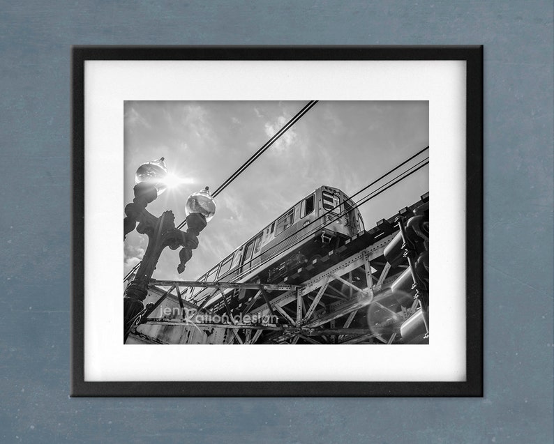 May include: A black and white photograph of a train passing under a bridge. The train is seen from a low angle, and the bridge is in the background. The train is partially obscured by the bridge, and the sun is shining brightly in the sky.
