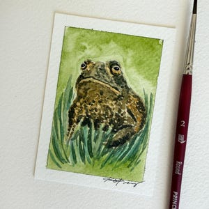 May include: A watercolor painting on paper depicting a textured brown toad sitting in green grass. The toad has dark spots and prominent eyes. A thin paintbrush with a maroon handle and the text "2 Round" is positioned vertically to the right.