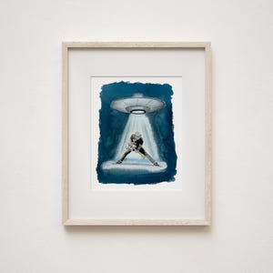 May include: Watercolor art print of a musician playing a guitar under a UFO's spotlight. The artwork features a blue and white color scheme, with the musician in a hat and dark clothing. The print is framed in a light wood frame.