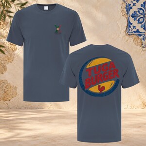 Könnte beinhalten: Ein schieferblaues Kurzarm-T-Shirt mit einem kleinen Logo auf der Brust, das den Buchstaben X und die portugiesische Flagge zeigt. Auf der Rückseite befindet sich eine große kreisförmige Grafik mit dem roten Text TUGA BURGER und einem roten Hahn.