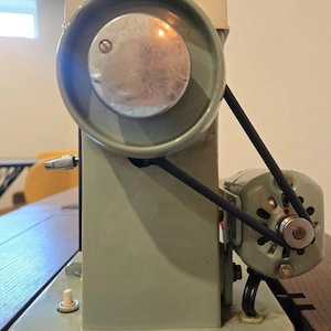 May include: A light sage green sewing machine side profile featuring a large circular handwheel with a metallic centre, a black rubber drive belt connecting to a small external motor, and a small white power switch on the base plate.