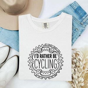 May include: A white short-sleeved t-shirt featuring a black graphic of a bicycle chainring and gear teeth. The text "I'D RATHER BE CYCLING" is printed in the center of the gear design. The shirt has a crew neckline and a Comfort Colors brand label.