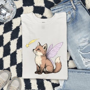 May include: A white t-shirt featuring a graphic illustration of a brown fox with light purple fairy wings, looking up at a small yellow shooting star. The fox has a white chest and a white-tipped tail, rendered in a clean, detailed line art style.