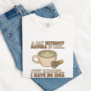 May include: A white t-shirt featuring a graphic of a beige ceramic cup filled with green matcha tea and a bamboo whisk. The text reads A DAY WITHOUT MATCHA IS LIKE... JUST KIDDING... I HAVE NO IDEA in a brown, retro-style font on the front.