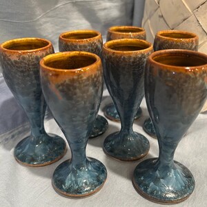 May include: A set of seven ceramic goblets featuring a crystalline glaze with deep blue and charcoal gray tones, accented by a warm amber rim. Each goblet has a smooth, tapered stem and a wide circular base, showcasing a unique, mottled texture.