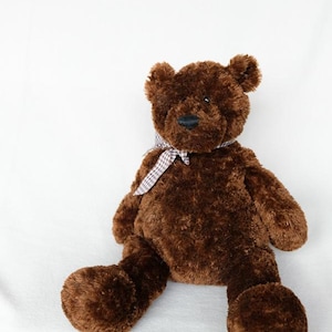 May include: A plush teddy bear with dark brown, textured fur, small rounded ears, and a black nose. The bear is wearing a small, brown and white chequered fabric ribbon tied in a bow around its neck, with two arms and two legs extended forward.