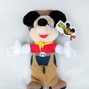 May include: A plush toy of a cartoon mouse dressed as a cowboy, wearing a brown hat, red shirt, yellow neckerchief, blue pants, and tan chaps. The toy has white gloves, black ears, and a tag attached to the ear that reads MOUSEKETOYS.