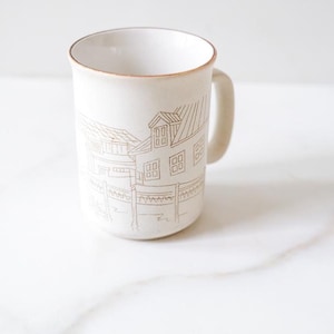 May include: A cream-coloured ceramic mug featuring a brown line-art illustration of a house with a fence. The mug has a smooth finish, a cylindrical body, a curved handle on the right side, and a thin brown rim around the top edge of the vessel.