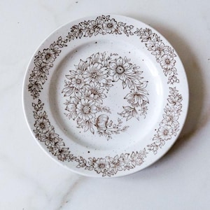 May include: A round white ceramic plate featuring a speckled finish and a brown line-art floral pattern. The design includes a continuous wreath of flowers around the rim and a central cluster of various blossoms and leaves in the middle.