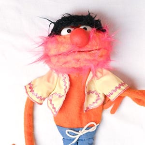 Op de afbeelding: Een handpop met een ronde rode neus, zwart haar en felroze vacht rond de mond. De pop draagt een crèmekleurig vestje met roze rand over een oranje lichaam en een blauwe broek met een wit trekkoord in de taille.