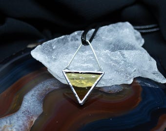 Puede incluir: Un colgante triangular con una pieza de vidrio texturizado de color amarillo pálido enmarcada en metal plateado. El colgante cuelga de un cordón negro fino y presenta un diseño geométrico con una sección superior angular y una base sólida.
