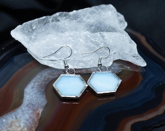 Puede incluir: Un par de pendientes colgantes con dijes de vidrio de color azul claro en forma hexagonal, engastados en un marco de metal plateado. Los pendientes están sujetos a ganchos de metal plateado para orejas perforadas.