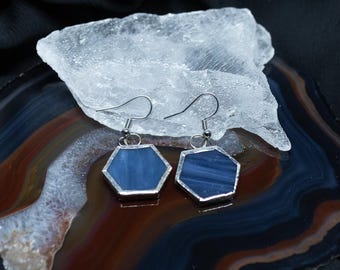 Puede incluir: Un par de pendientes colgantes con piezas de vidrio hexagonales de color azul oscuro engastadas en marcos de metal plateado. Cada pendiente cuelga de un gancho sencillo, mostrando una superficie lisa y de color sólido en su forma geométrica.