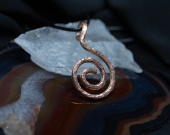 Puede incluir: Un colgante de metal color cobre con forma de espiral y una superficie martillada y texturizada. El colgante está suspendido de un cordón negro fino y presenta un diseño en espiral distintivo que termina en un bucle para el cordón.