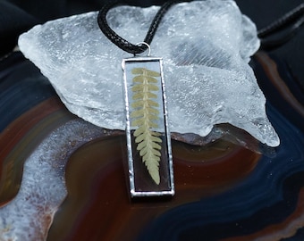 Puede incluir: Un colgante rectangular que contiene una hoja de helecho verde prensada en resina transparente con un marco de metal plateado. El colgante cuelga de un cordón negro, mostrando un diseño botánico que resalta la textura natural del helecho.