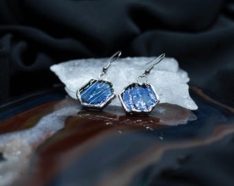 Puede incluir: Un par de pendientes hexagonales con superficies texturizadas en azul profundo, enmarcadas en metal de tono plateado. Cada pendiente cuelga de un fino gancho plateado, mostrando un patrón distintivo de líneas blancas y tonos azul oscuro.