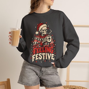 May include: A dark gray crewneck sweatshirt featuring a graphic of a skeleton wearing a Santa hat and a patterned holiday sweater. The skeleton holds a mug with a skull icon. The text reads FEELING FESTIVE in red and white, surrounded by Christmas lights.