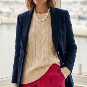 May include: A woman wearing a navy blue blazer over a cream-colored cable-knit sweater and bright pink corduroy pants. The pants have a repeating dark pattern. She is also wearing a pearl necklace and earrings.