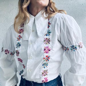 Op de afbeelding: Witte blouse met kleurrijke bloemenborduursels op de voorkant en mouwen. De blouse heeft een brede kraag, een knoopsluiting en pofmouwen. De borduursels bevatten tinten roze, blauw, groen en geel.