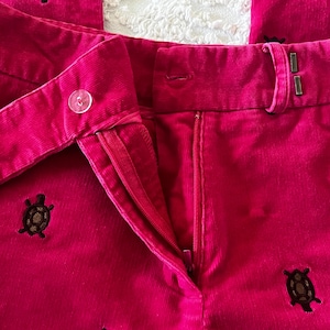 May include: Close-up of bright pink corduroy pants with a button and zipper closure. The pants feature embroidered brown turtle designs scattered across the fabric. The pants have a classic, tailored style.