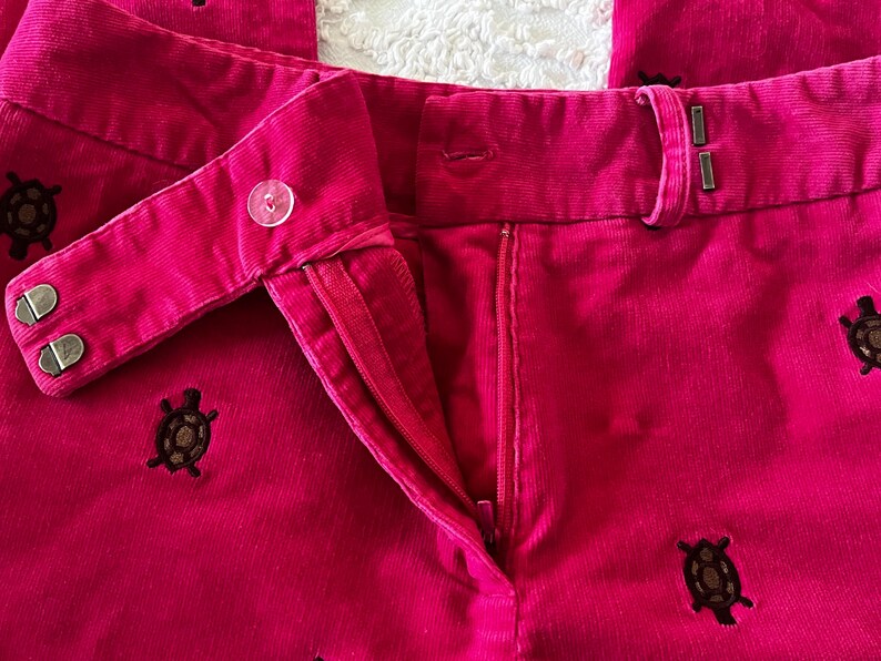 May include: Close-up of bright pink corduroy pants with a button and zipper closure. The pants feature embroidered brown turtle designs scattered across the fabric. The pants have a classic, tailored style.