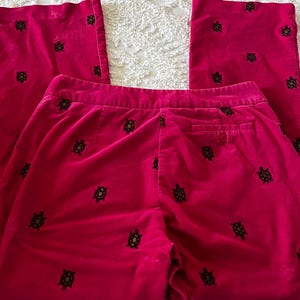 May include: Bright pink corduroy pants with a wide leg. The pants are adorned with a pattern of small, dark green turtles. The pants have a button closure and a back pocket. The fabric has a soft, velvety texture.