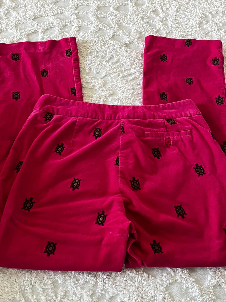 May include: Bright pink corduroy pants with a wide leg. The pants are adorned with a pattern of small, dark green turtles. The pants have a button closure and a back pocket. The fabric has a soft, velvety texture.