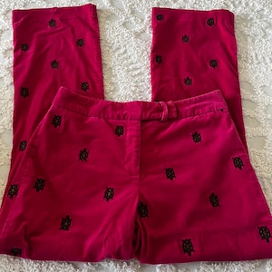May include: Bright pink velvet pants with a straight leg cut. The pants are adorned with a pattern of small, embroidered turtles in black and gold. The pants have a button and zipper closure and belt loops.